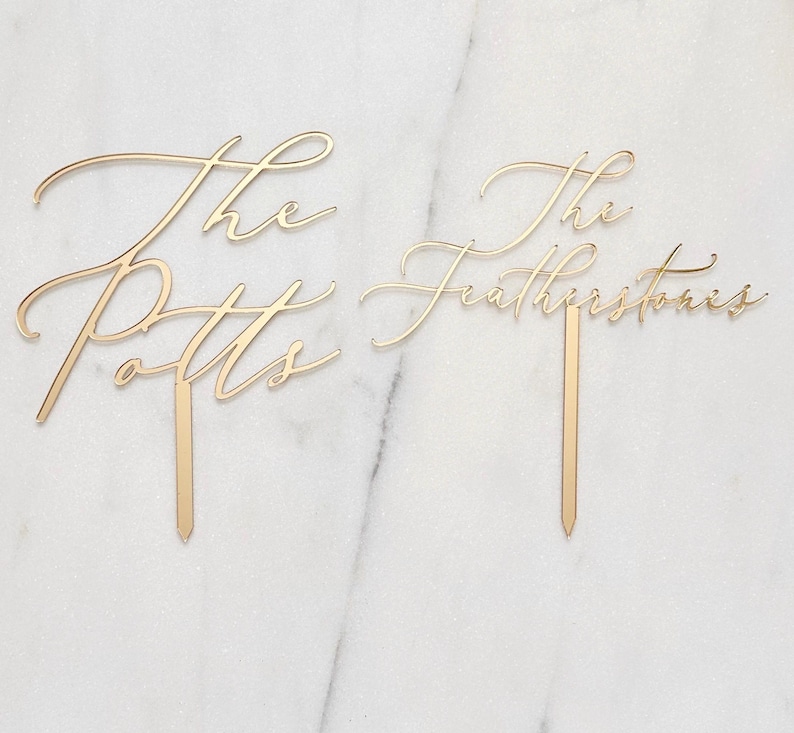 K&ouml;nnte beinhalten: Zwei goldfarbene Tortenaufs&auml;tze mit eleganter Schreibschrift. Einer tr&auml;gt die Aufschrift "The Potts" und der andere "The Featherstones". Jeder Aufsatz hat einen schmalen, spitzen Stab zum Einstecken in einen Kuchen.