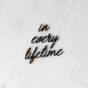 May include: Black cursive text on a white background reads "in every lifetime." The text is in a flowing script style, with the letters connected. The background is a textured, off-white surface.