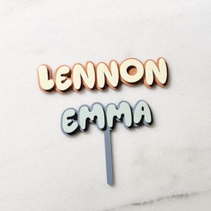 Op de afbeelding: Twee gepersonaliseerde naamtoppers. Het woord "LENNON" is in lichtbruin en crème. Het woord "EMMA" is in lichtblauw en wit met een grijze stok.