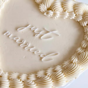 Op de afbeelding: Een hartvormige taart met witte glazuur en spuitroomranden. De woorden "just married" zijn in elegant wit schrift over de bovenkant geschreven. Een feestelijk dessert voor een bruiloft of jubileum.