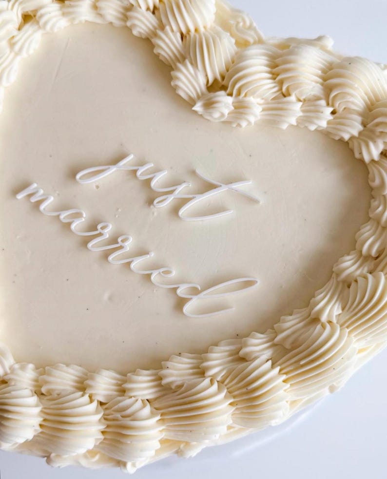 Puede incluir: Un pastel blanco en forma de coraz&oacute;n con glaseado blanco de crema de mantequilla y las palabras "Just Married" escritas en glaseado blanco.