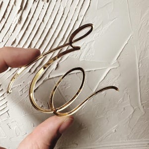 Op de afbeelding: Een goudkleurige acryl letter E in een cursief lettertype. De letter heeft een reflecterende, metallic afwerking met gladde, gebogen lijnen en een gepolijst oppervlak. Het ontwerp is lichtgewicht en uitgesneden in een gestileerde, elegante kalligrafische vorm.