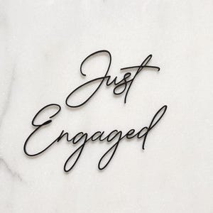 Op de afbeelding: Zwarte cursieve tekst "Just Engaged" op een witte marmeren achtergrond. Het elegante schrift is een decoratief element, perfect voor verlovingsaankondigingen of huwelijksdecoratie.