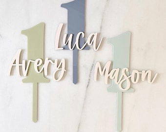 May include: Three cake toppers featuring the number one in sage green, slate blue, and mint green. Each topper includes a white script name cutout: Avery, Luca, and Mason. The toppers have a pointed stake at the bottom for insertion.