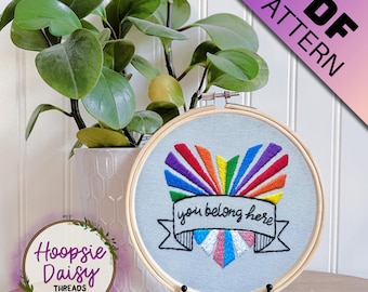 May include: Embroidered hoop art with a rainbow heart and the text "you belong here". The heart is surrounded by a banner with a rainbow and transgender pride flag.