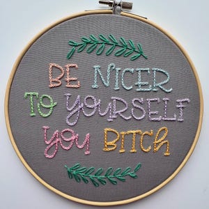 May include: A gray fabric embroidery hoop with the phrase "BE NICER TO YOURSELF YOU BITCH" stitched in colorful thread. Green leaf accents are stitched at the top and bottom of the hoop. The wooden hoop is visible.