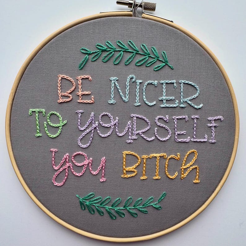 May include: A gray fabric embroidery hoop with the phrase "BE NICER TO YOURSELF YOU BITCH" stitched in colorful thread. Green leaf accents are stitched at the top and bottom of the hoop. The wooden hoop is visible.