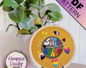 May include: An embroidered hoop featuring the words "SAFE SPACE" in rainbow hues, encircled by hearts and a planet design. The hoop is displayed on a black stand, with a plant in a white pot in the background. The text "PDF PATTERN" is in the upper right corner.