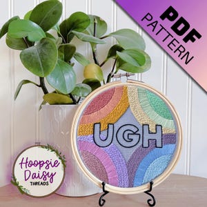 May include: Embroidered hoop art with the word "UGH" in black letters on a gray background. The background is filled with colorful rainbow-like arcs of embroidery floss.