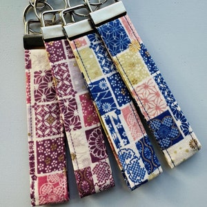 May include: Four keychains with different patterned fabric straps. The straps are in shades of pink, white, blue, and gold. The keychains are attached to silver key rings.