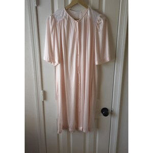 May include: A pale pink knee-length robe featuring short sleeves, a button-down front closure, and white lace detailing across the shoulder panels. The garment is made of a smooth, lightweight fabric with a slight sheen and a gathered neckline.