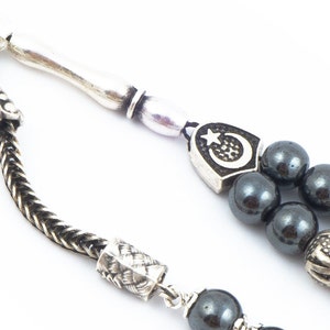 May include: A silver and black prayer beads with a crescent moon and star charm. The beads are made of hematite and the charm is silver.