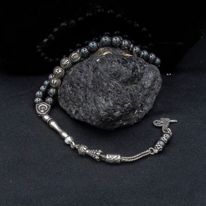May include: A black hematite prayer bead mala with silver accents and a silver tassel. The beads are arranged in a circle on a dark surface.