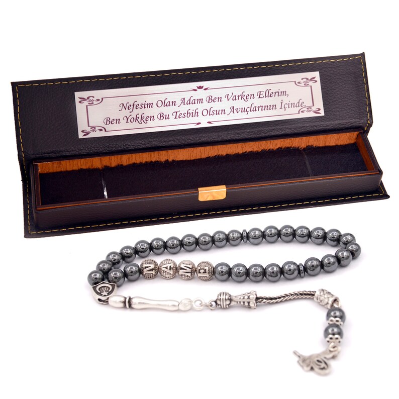 May include: A black leather box with a silver and black hematite prayer beads. The box has a quote in Turkish that reads: "Nefesim Olan Adam Ben Varken Ellerim, Ben Yokken Bu Tesbih Olsun Avu&ccedil;larının İ&ccedil;inde."