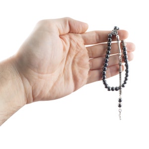 May include: A black beaded prayer chain with a silver charm and a silver tassel. The chain is held in a hand.