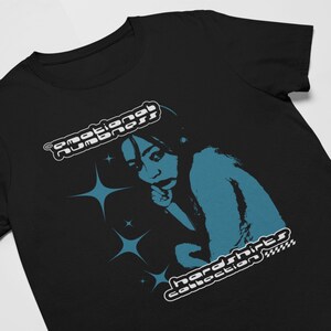 May include: Black t-shirt featuring a teal graphic of a person with a finger to their lips, surrounded by star shapes. The shirt has the text "emotional numbness" and "hardshirts collection" in a retro font.
