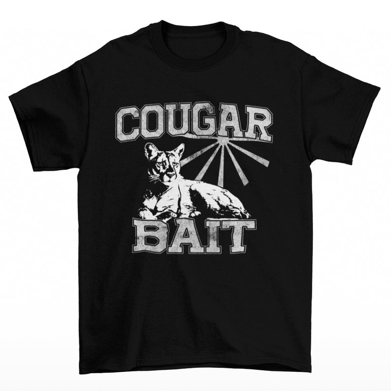 May include: Black t-shirt with a distressed white graphic. The graphic features the words "COUGAR" above and "BAIT" below a stylized image of a cougar. The cougar is lying down with a sunburst design behind it.