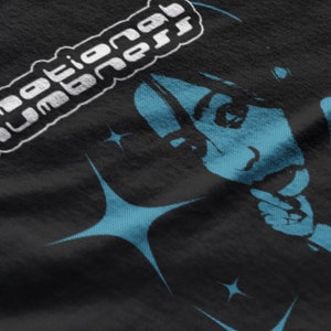 May include: Black t-shirt featuring a stylized illustration of a person with blue accents and starburst shapes. The text "emotional numbness" is printed in white, in a rounded, stylized font.