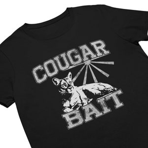 May include: Black t-shirt with a distressed graphic design. The design features the words "COUGAR BAIT" in a vintage, collegiate-style font, with an illustration of a cougar. The shirt is made of a soft material.