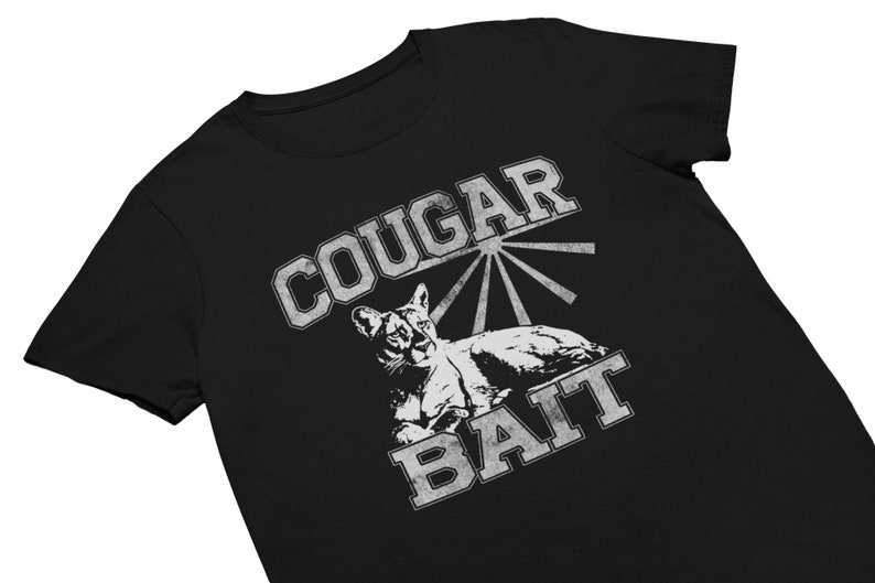 May include: Black t-shirt with a distressed graphic design. The design features the words "COUGAR BAIT" in a vintage, collegiate-style font, with an illustration of a cougar. The shirt is made of a soft material.