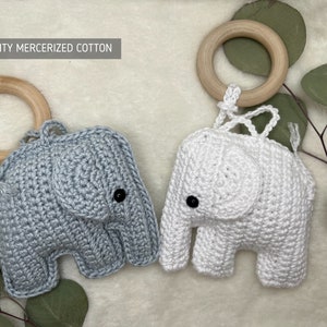 Pu&ograve; includere: Due giocattoli a forma di elefante all'uncinetto, uno grigio e uno bianco, ciascuno con un anello di legno e occhi a bottone neri. Il testo "HIGH QUALITY MERCERIZED COTTON" &egrave; visibile. I giocattoli sono su uno sfondo morbido e neutro con fogliame verde.