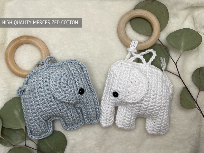 Pu&ograve; includere: Due giocattoli a forma di elefante all'uncinetto, uno grigio e uno bianco, ciascuno con un anello di legno e occhi a bottone neri. Il testo "HIGH QUALITY MERCERIZED COTTON" &egrave; visibile. I giocattoli sono su uno sfondo morbido e neutro con fogliame verde.