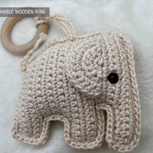 Pu&ograve; includere: Un giocattolo per bambini a forma di elefante all'uncinetto, di colore beige chiaro. Presenta un occhio a bottone nero e un anello di legno staccabile. Il testo "DETACHABLE WOODEN RING" &egrave; visibile.