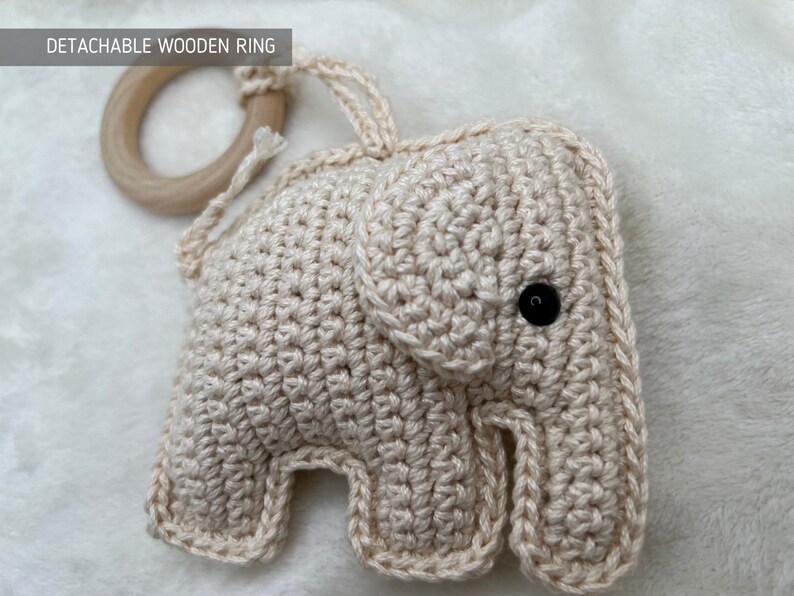 Pu&ograve; includere: Un giocattolo per bambini a forma di elefante all'uncinetto, di colore beige chiaro. Presenta un occhio a bottone nero e un anello di legno staccabile. Il testo "DETACHABLE WOODEN RING" &egrave; visibile.