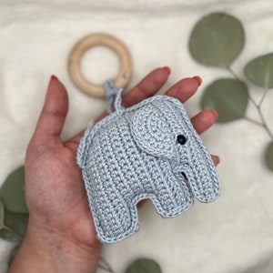 Pu&ograve; includere: Un giocattolo a forma di elefante all'uncinetto grigio chiaro con un anello di legno. L'elefante ha occhi neri ed &egrave; tenuto in mano. Il giocattolo &egrave; progettato per neonati e bambini piccoli.
