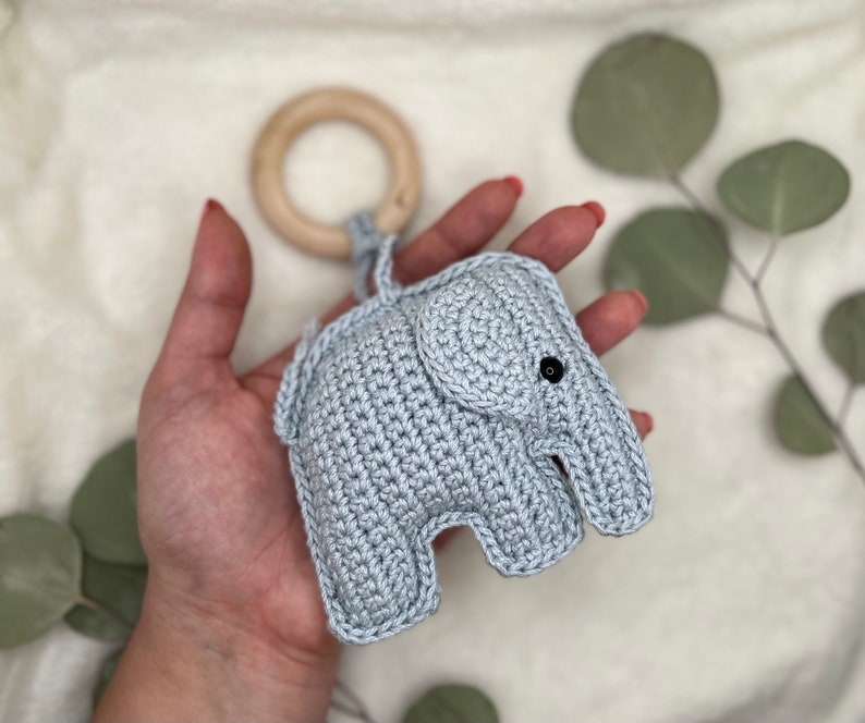 Pu&ograve; includere: Un giocattolo a forma di elefante all'uncinetto grigio chiaro con un anello di legno. L'elefante ha occhi neri ed &egrave; tenuto in mano. Il giocattolo &egrave; progettato per neonati e bambini piccoli.
