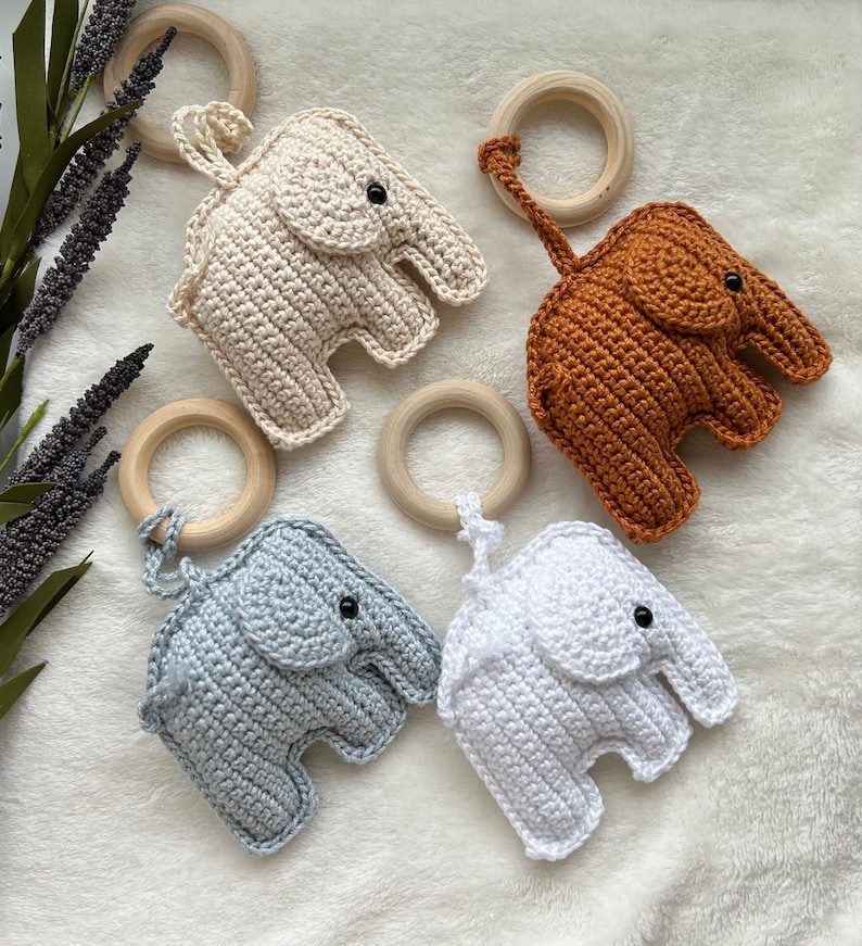 Pu&ograve; includere: Quattro giocattoli a forma di elefante all'uncinetto in crema, marrone, azzurro e bianco, ciascuno attaccato a un anello di legno. Gli elefanti hanno occhi neri a bottone e sono disposti su una superficie bianca e morbida. I giocattoli sono fatti a mano.
