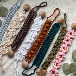 May include: A collection of handmade crochet pacifier clips in various colors, including blue, beige, brown, white, orange, green, and pink. Each clip features a wooden bead and a metal clip for attachment. The designs include textured patterns and floral accents.
