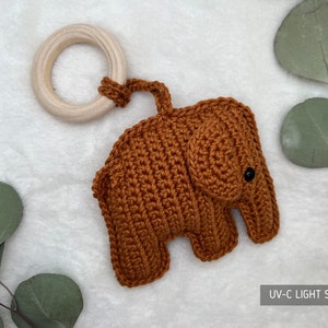 Pu&ograve; includere: Un giocattolo per bambini a forma di elefante all'uncinetto, in una calda tonalit&agrave; marrone, attaccato a un anello di legno naturale. Il giocattolo presenta una superficie strutturata e un piccolo occhio nero. Il testo "UV-C LIGHT SANITIZED" &egrave; visibile.