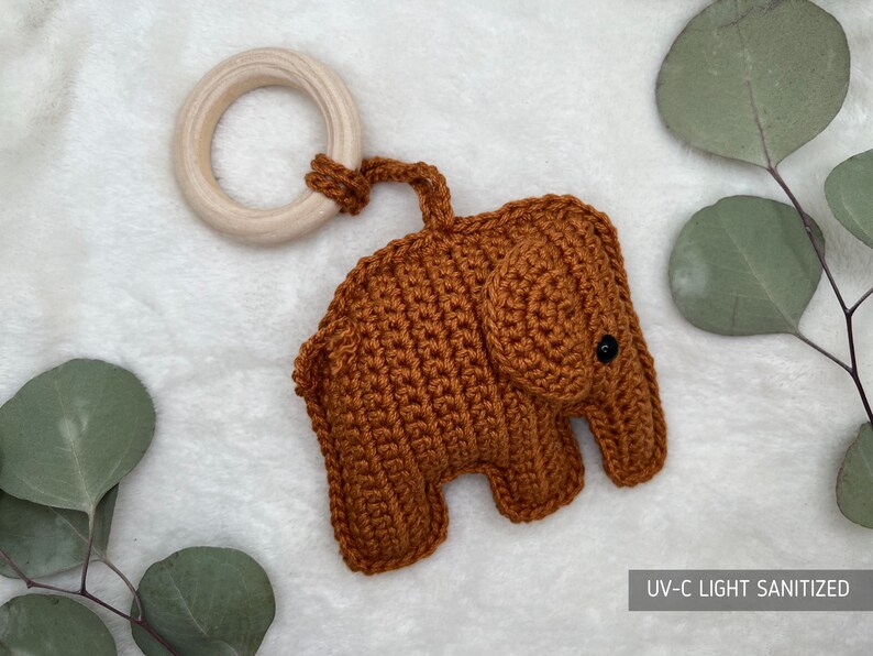 Pu&ograve; includere: Un giocattolo per bambini a forma di elefante all'uncinetto, in una calda tonalit&agrave; marrone, attaccato a un anello di legno naturale. Il giocattolo presenta una superficie strutturata e un piccolo occhio nero. Il testo "UV-C LIGHT SANITIZED" &egrave; visibile.
