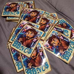 以下が含まれることがあります： メガネをかけた男性の漫画イラストと「BBNC$」の文字が入った長方形のステッカーのコレクション。ステッカーは金色の縁取りと青とクリーム色の配色です。