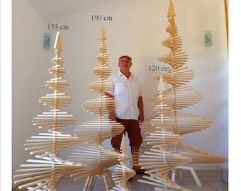 May include: Four wooden Christmas trees of varying sizes, standing on a tiled floor. The trees are made of thin wooden slats arranged in a spiral pattern. The tallest tree is 240 cm tall, the next tallest is 190 cm, the next is 155 cm, and the smallest is 65 cm tall. A person is standing next to the trees, providing a scale reference.