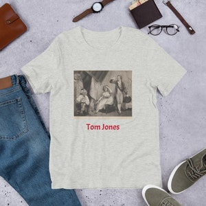 Puede incluir: Una camiseta gris jaspeada con una ilustración vintage de una escena de la novela "Tom Jones" de Henry Fielding. La ilustración muestra a cuatro personas en una habitación, con una mujer sentada en una silla y un hombre de pie junto a ella. El texto "Tom Jones" está impreso debajo de la ilustración.