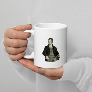 May include: White ceramic mug featuring a black and white image of a man in a suit and tie. The man is seated in a chair and holding a book.