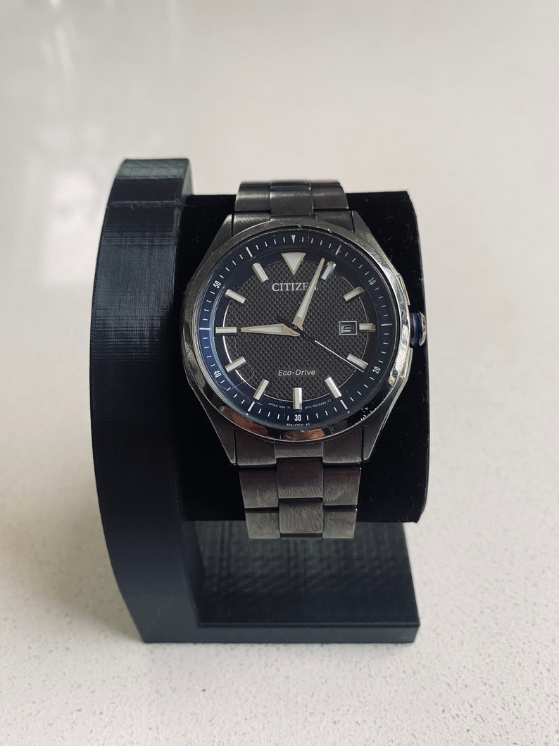 May include: A black Citizen Eco-Drive watch with a silver stainless steel band. The watch face is black with silver hands and markers. The watch is on a black watch stand.