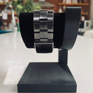 May include: A black watch stand with a textured base and a curved arm to hold a watch. The watch has a silver metal band.