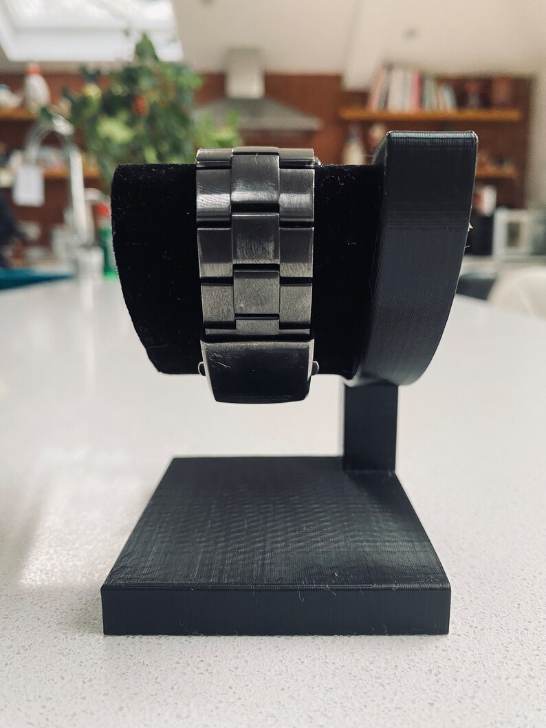 May include: A black watch stand with a textured base and a curved arm to hold a watch. The watch has a silver metal band.