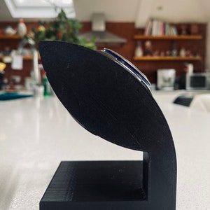 May include: A black 3D printed stand for holding a watch. The stand is shaped like a curved, open-ended cylinder with a rectangular base.