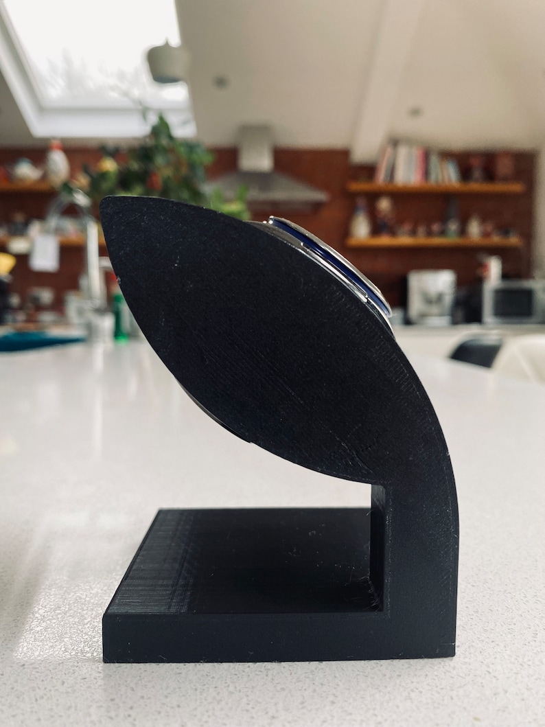 May include: A black 3D printed stand for holding a watch. The stand is shaped like a curved, open-ended cylinder with a rectangular base.