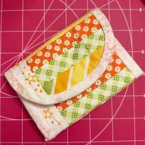 May include: A handmade, quilted wallet with a flap closure. The wallet features a patchwork design with orange floral, green gingham, and yellow striped fabrics. The edges are trimmed with a white fabric and embroidered with orange stars. The wallet is approximately 5 inches wide and 4 inches tall.