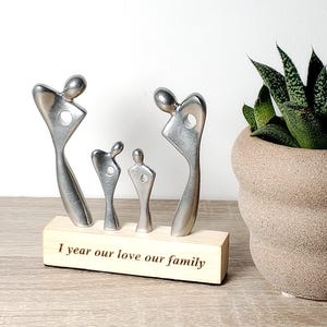 Może przedstawiać: Metalowa rzeźba rodziny z czterema figurami na drewnianej podstawie. Na podstawie wygrawerowano napis "I year our love our family". Po prawej stronie znajduje się roślina doniczkowa.