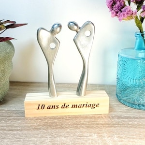 May include: A silver metal sculpture of two abstract figures facing each other, mounted on a wooden base. The base is engraved with the text "10 ans de mariage". A blue glass vase with purple flowers is in the background.