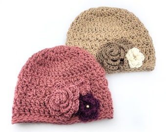 May include: Two crocheted hats with flower embellishments. One hat is a light brown color with a brown and white flower. The other hat is a pink color with a pink and purple flower.