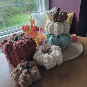 May include: Five crocheted pumpkins in various shades of brown, white, and blue. The pumpkins are arranged on a wooden table with a fall-themed background.