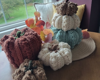 May include: Five crocheted pumpkins in various shades of brown, white, and blue. The pumpkins are arranged on a wooden table with a fall-themed background.