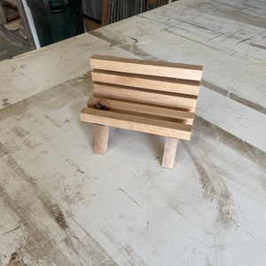 Può includere: Una piccola panchina di legno con quattro doghe e due gambe su ogni lato. La panchina è in legno marrone chiaro e si trova su una superficie bianca.